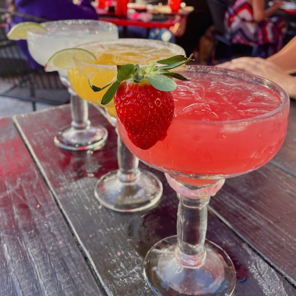 Bright pink cocktail garnished with a fresh strawberry, accompanied by citrus-garnished drinks on a restaurant table.