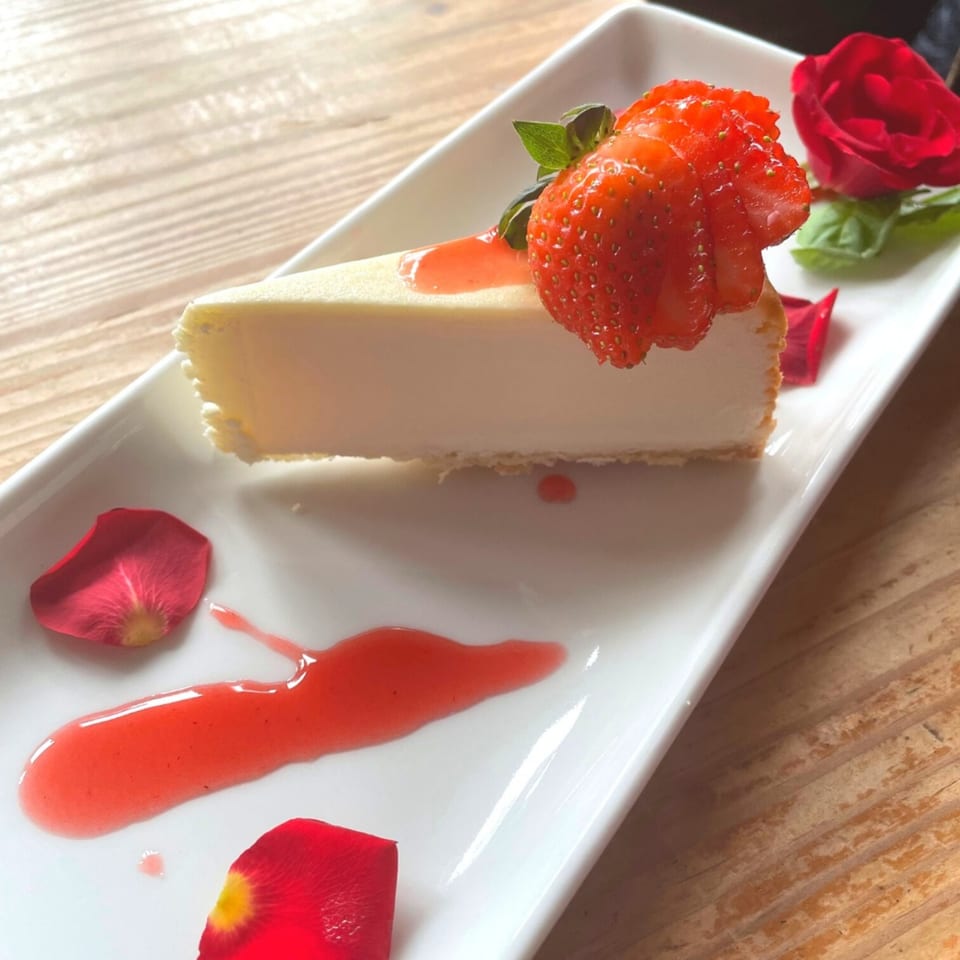 A slice of cheesecake with strawberry topping, strawberry drizzle, and rose petals on a white plate.
