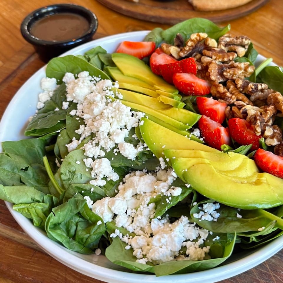 A vibrant spinach salad with strawberries, avocado, walnuts, and feta cheese, with dressing on the side.