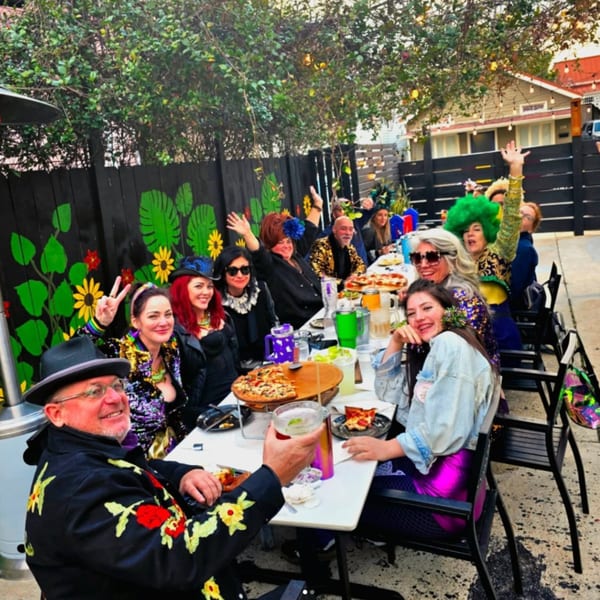 People in festive attire enjoying pizza and drinks at an outdoor restaurant table.