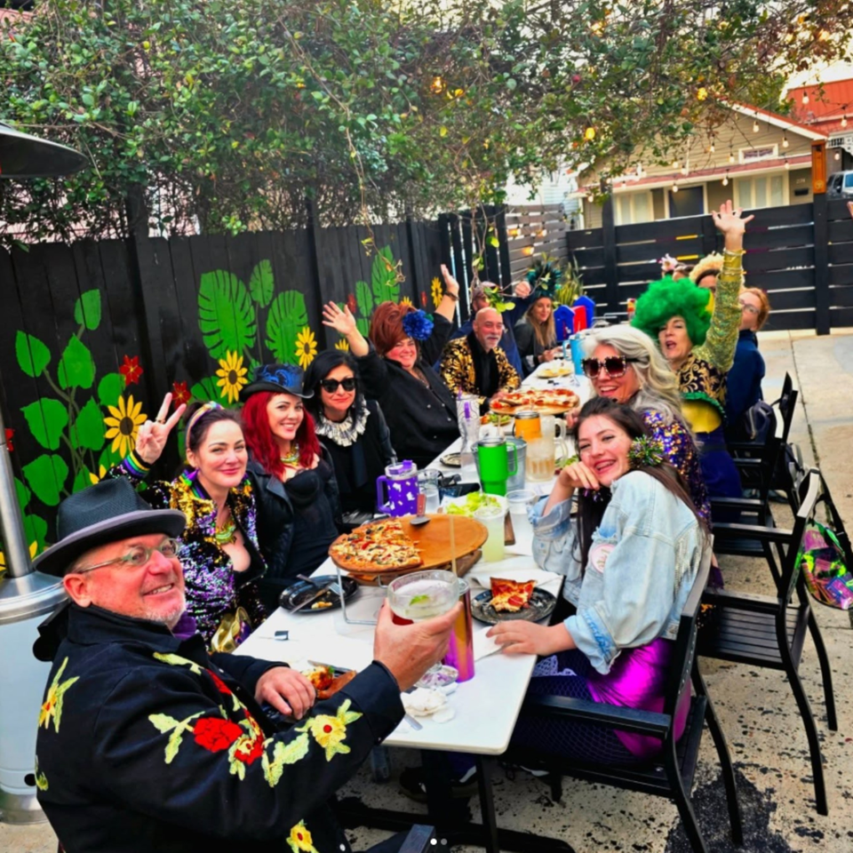 People in festive attire enjoying pizza and drinks at an outdoor restaurant table.