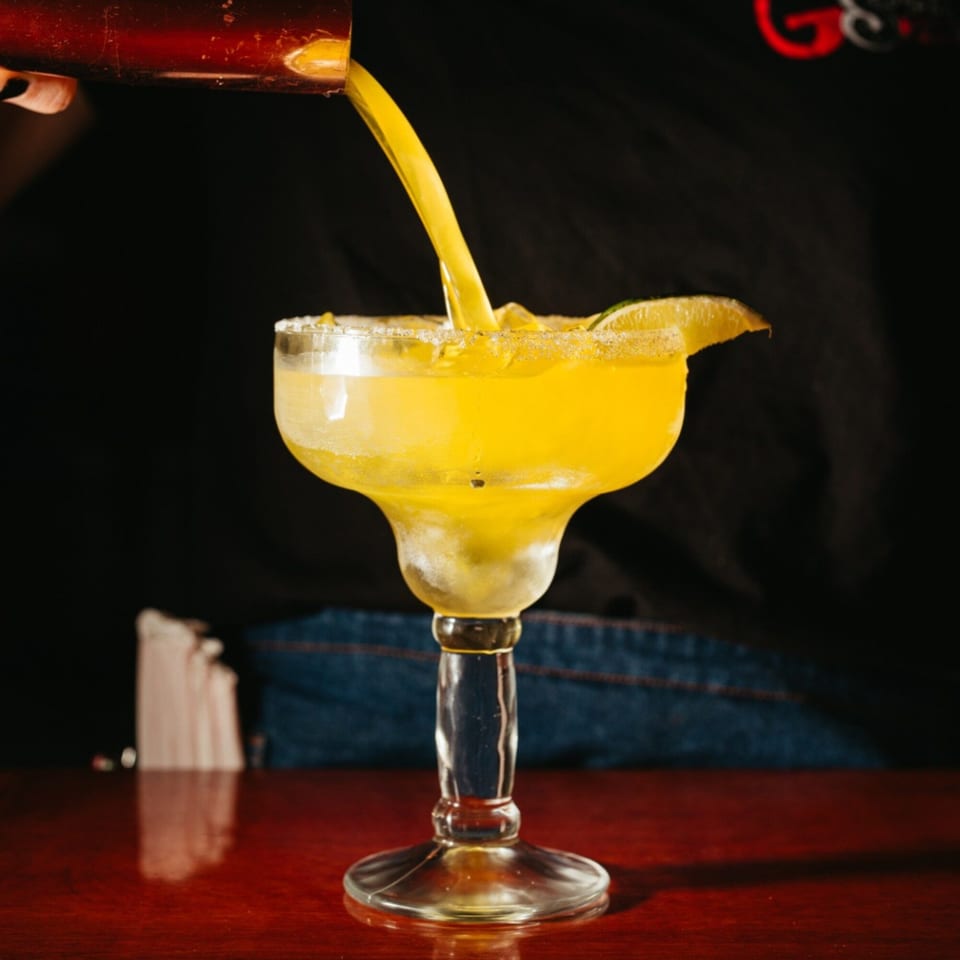 A bright yellow margarita is poured into a salt-rimmed glass with a lime wedge.