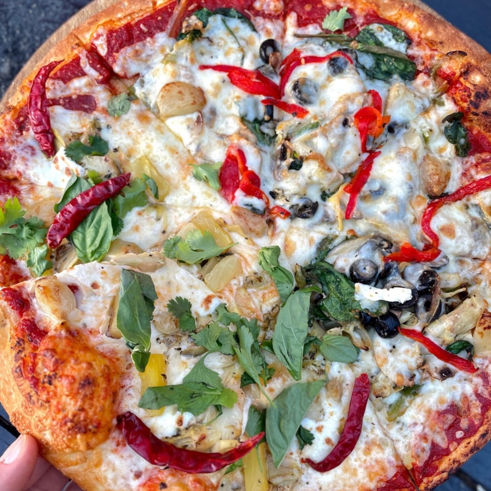 Close-up of a G's Pizza with various toppings including red peppers, olives, and herbs.