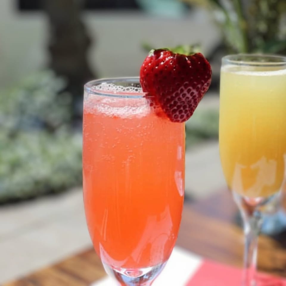 Bright pink strawberry drink garnished with a fresh strawberry beside a pale yellow beverage on a restaurant table.