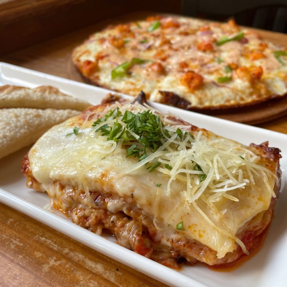 A cheesy lasagna with parsley and shredded cheese, flatbread, and a G's Pizza in the background.